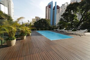 Piscina no terraço com espreguiçadeiras, plantas em vasos e edifícios altos ao fundo.