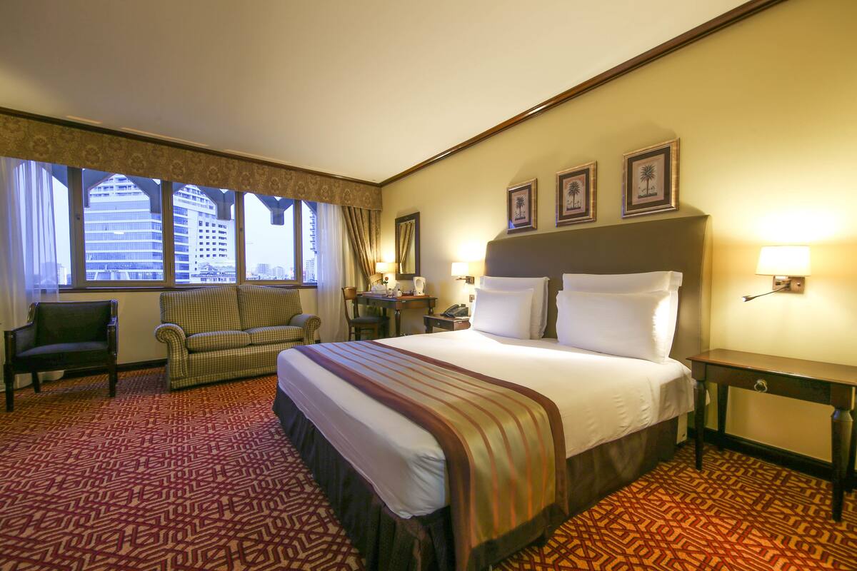 A hotel room with a large bed, a chair, a desk, and a view of the city through large windows.