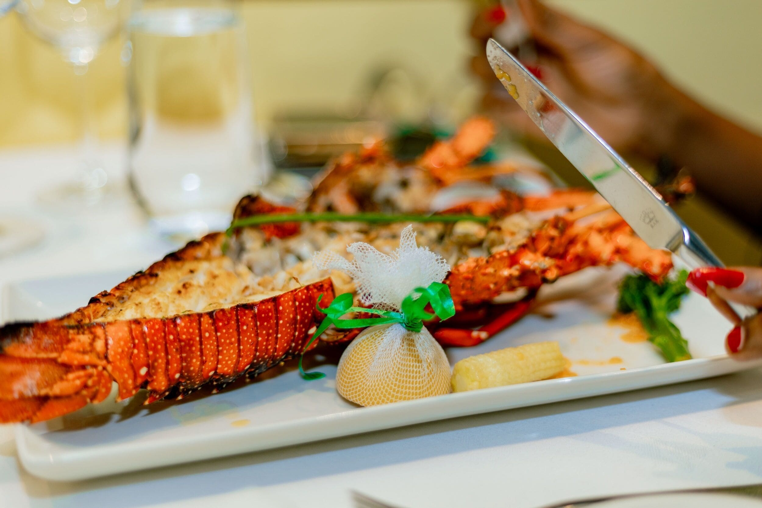 Une queue de homard servie sur une assiette blanche, décorée d’un ruban vert, et agrémentée d’une petite fleur blanche et d’une tranche de citron.
