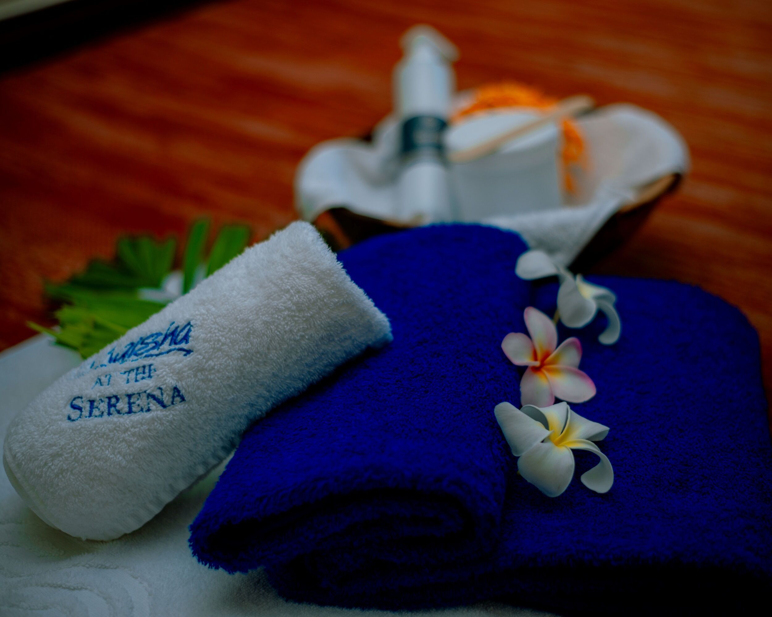 A towel with "Serena" embroidered on it, a blue towel, and a bottle of lotion on a wooden surface.
