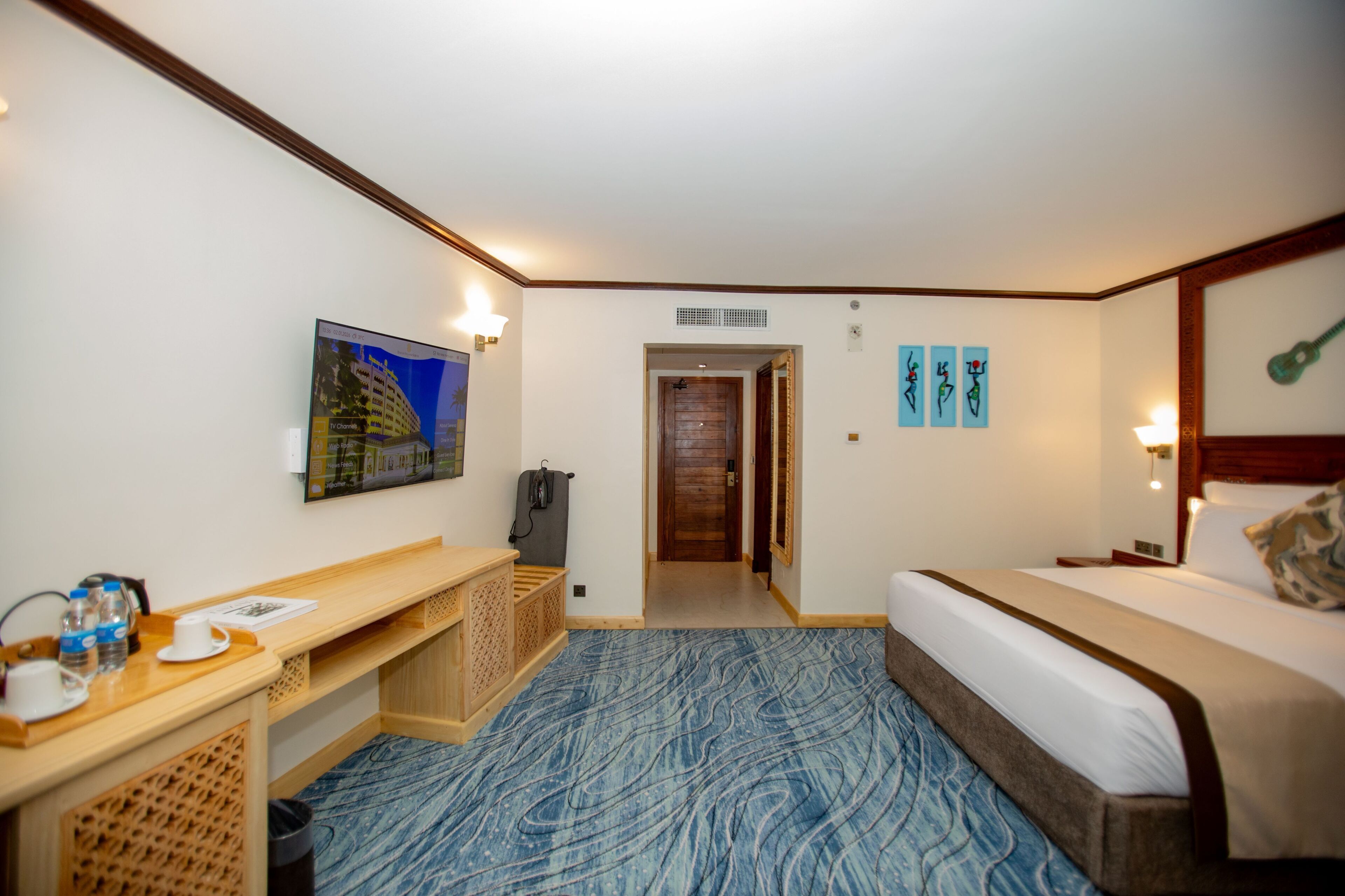 Une chambre d’hôtel comprenant un lit, un bureau avec une chaise, une petite table avec des bouteilles d’eau, un tableau au mur et une porte donnant sur une autre pièce.