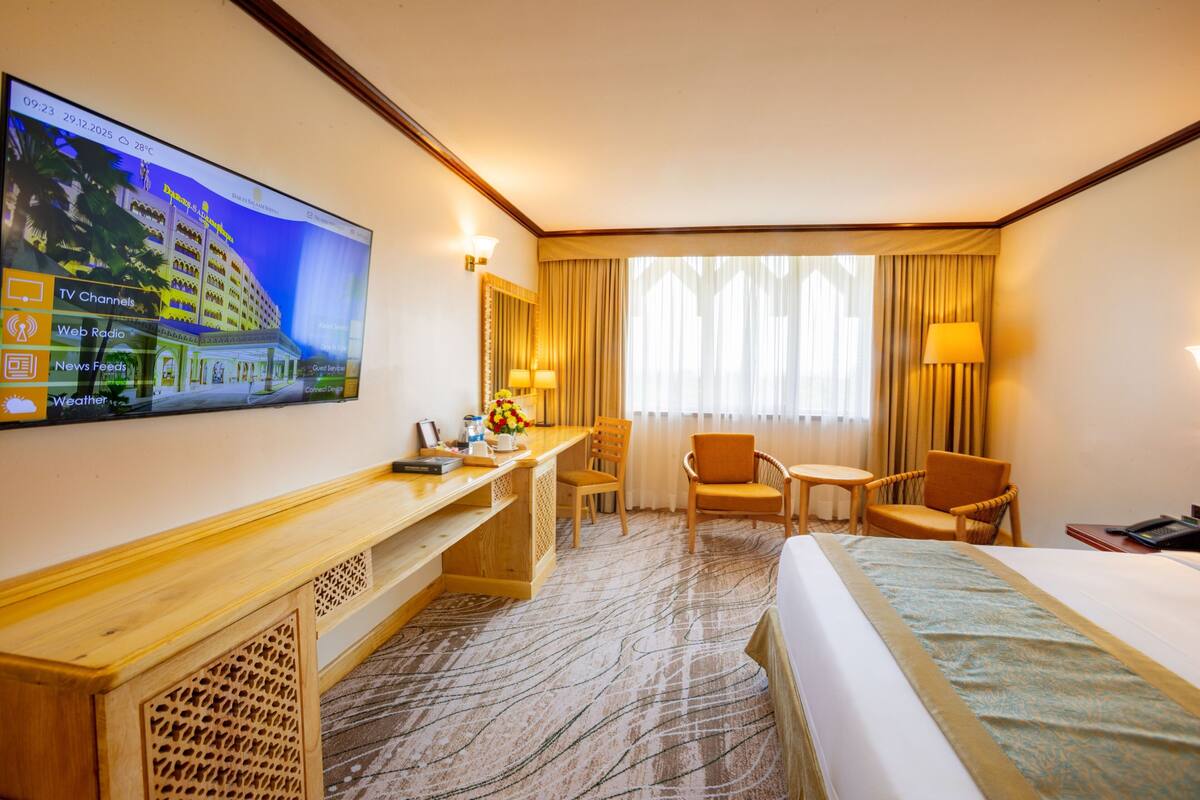 A hotel room with a bed, desk, chairs, a TV on the wall, and a window with curtains.