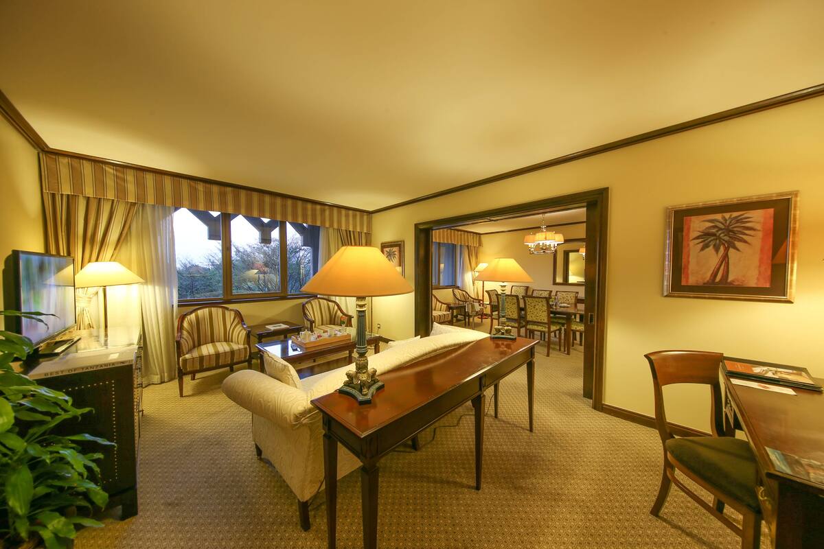 A hotel room with a large window, a dining table, two lamps, a sofa, and a chair.