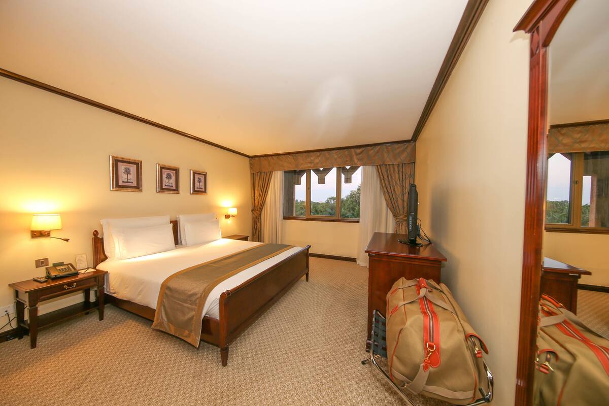 A hotel room with a large bed, bedside table with a lamp, a dresser, and a window with curtains.
