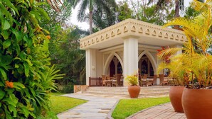 A covered outdoor seating area with a tiled floor, surrounded by lush greenery and palm trees. - Dar Es Salaam Serena Hotel (Dar es Salaam)