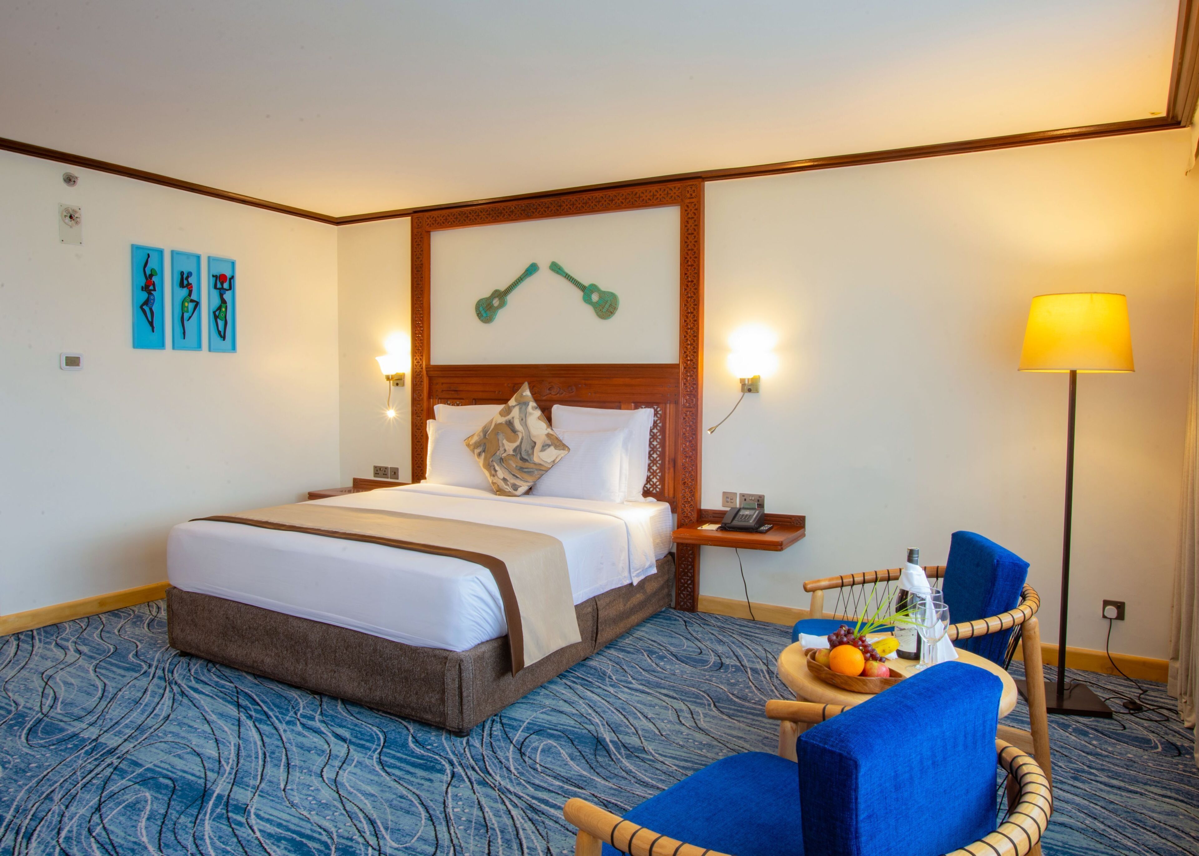 Ein Hotelzimmer mit einem großen Bett, einem Nachttisch mit Lampe, einem Sessel mit Obstschale und Bildern an den Wänden.