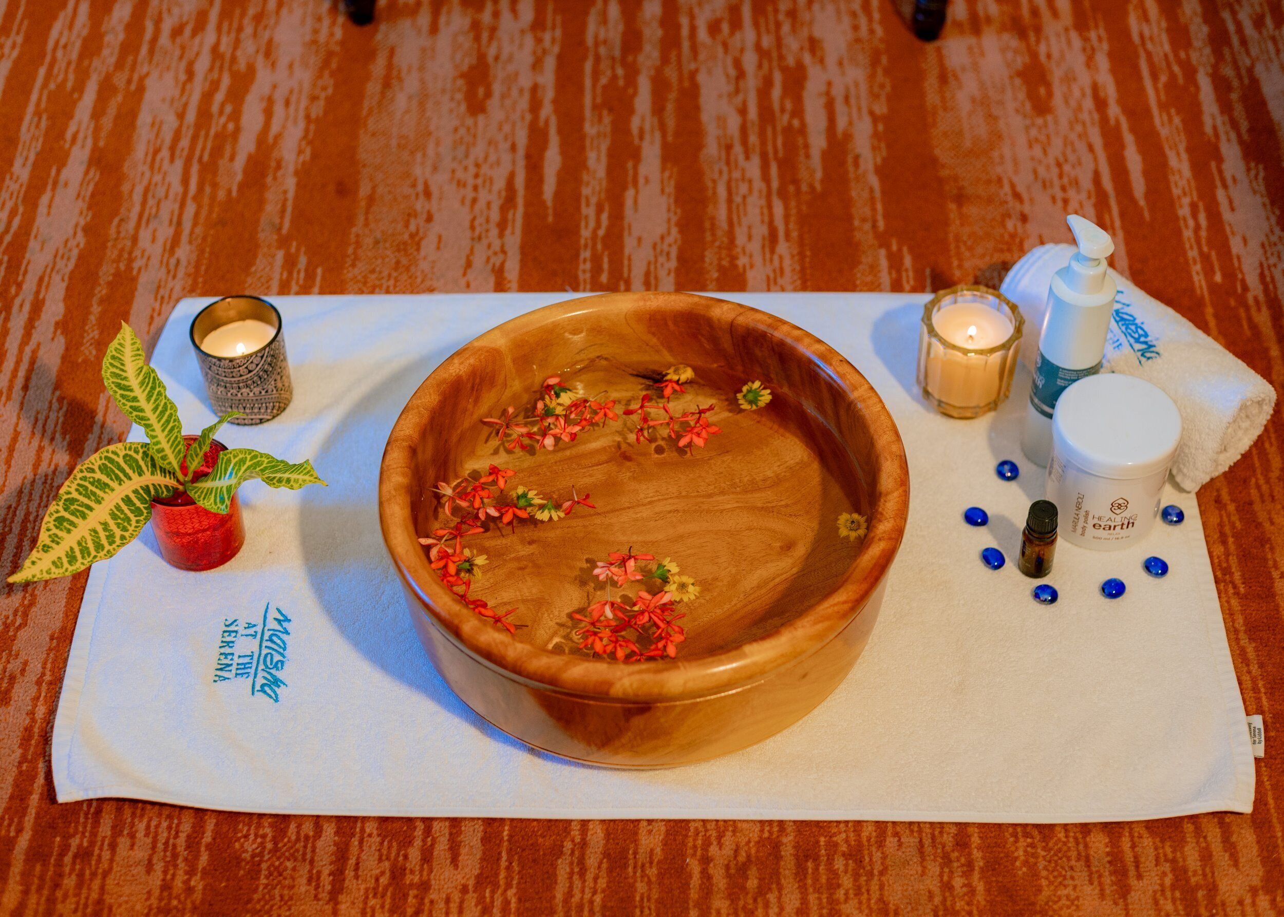 Eine Holzkasse mit Blumen auf weißem Stoff, eine kleine Topfpflanze, Kerzen und eine Lotionflasche auf einem Holzboden.