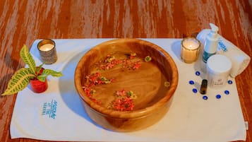 A wooden bowl with flowers on a white cloth, a small potted plant, candles, and a bottle of lotion on a wooden floor.
