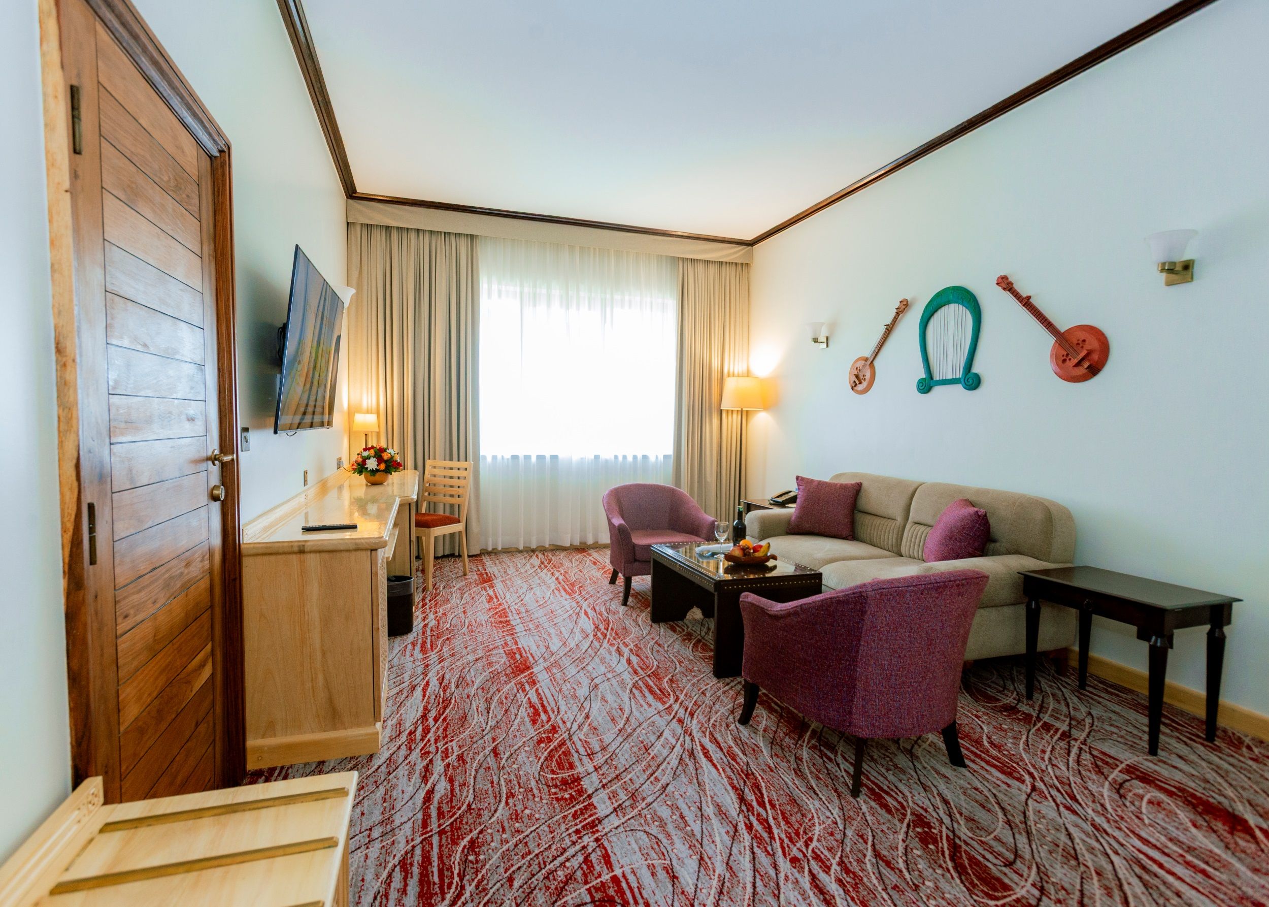 Ein Hotelzimmer mit Holztür, einem an der Wand montierten Fernseher, einem Essbereich mit Tisch und Stühlen, einer Couch, einem Couchtisch und einem gemusterten Teppich.