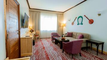 Ein Hotelzimmer mit Holztür, einem an der Wand montierten Fernseher, einem Essbereich mit Tisch und Stühlen, einer Couch, einem Couchtisch und einem gemusterten Teppich.