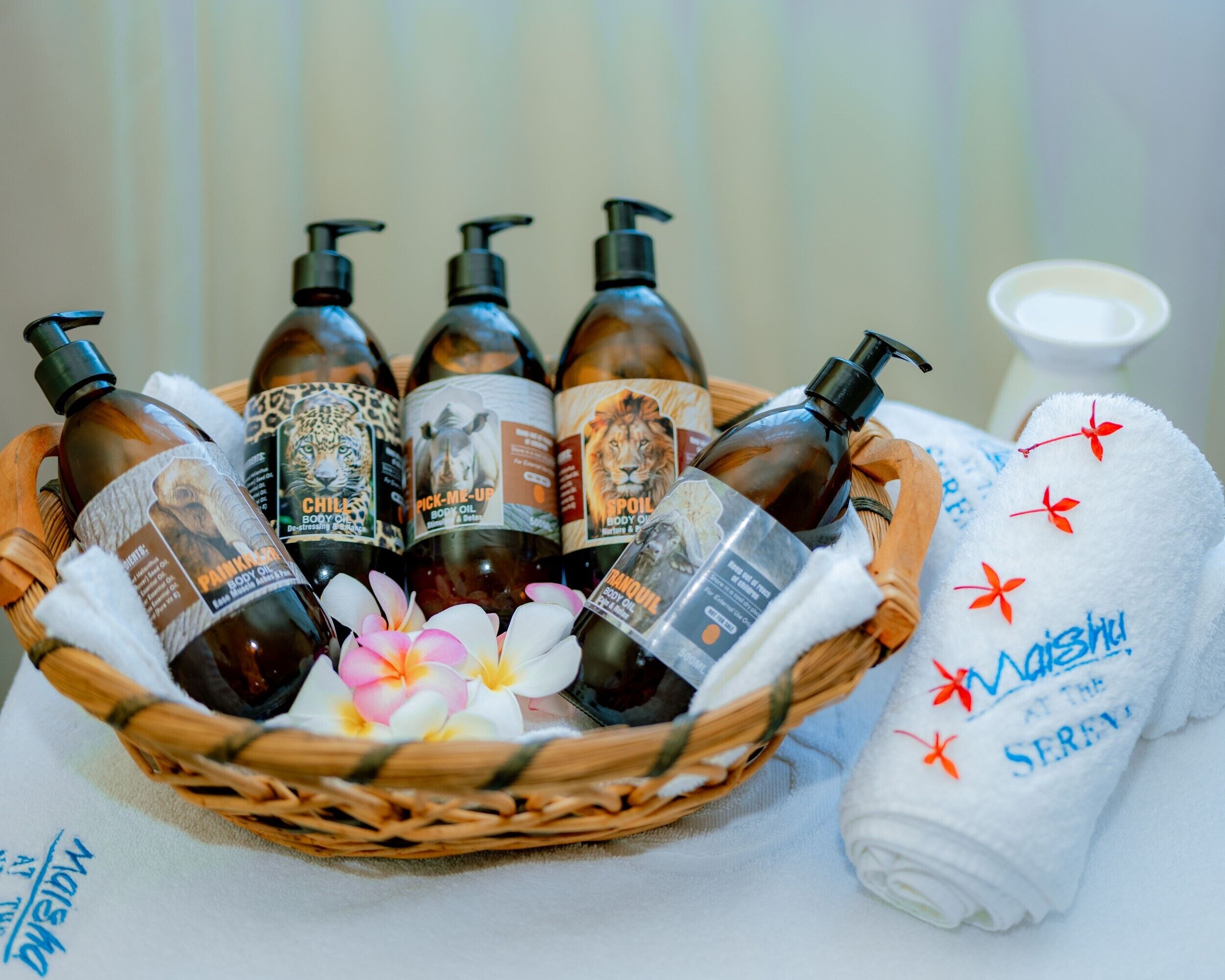 Una cesta con botellas de aceite corporal etiquetadas, una toalla blanca con texto bordado y una taza blanca.