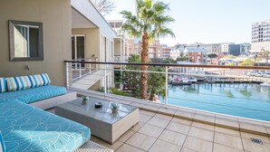 A balcony with a sofa and a coffee table, overlooking a marina with boats and buildings in the background. - Waterfront Village (Cape Town)