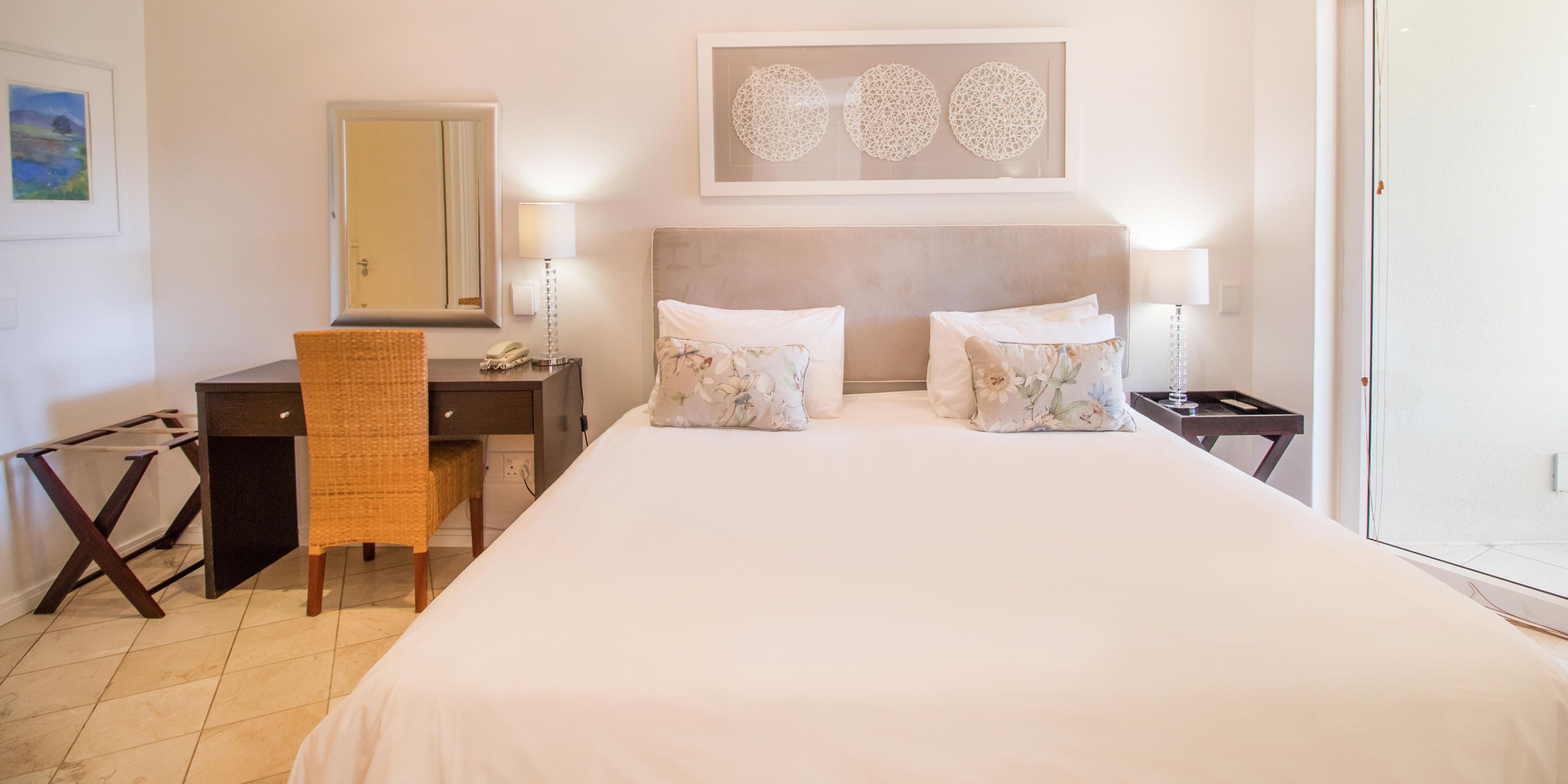 Une chambre d’hôtel comprenant un lit, deux tables de chevet, un bureau avec une chaise, un miroir et une œuvre d’art accrochée au mur.
