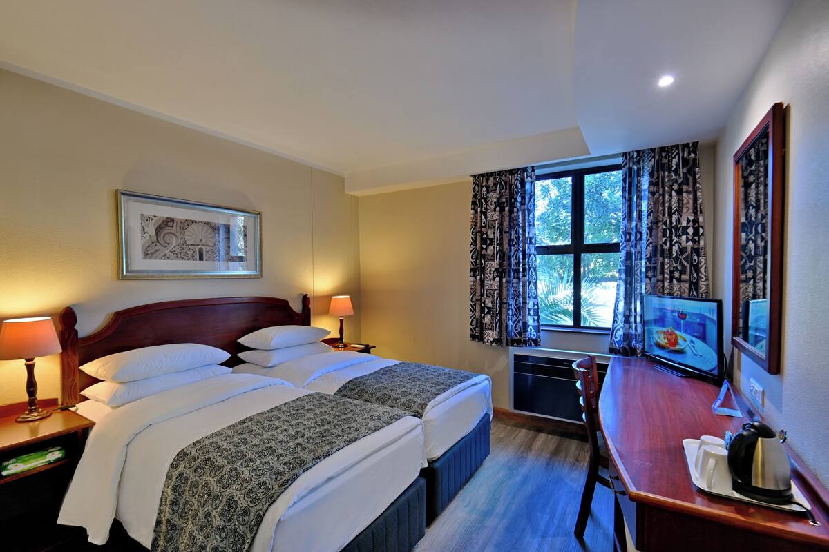 Une chambre d’hôtel avec un grand lit, deux tables de chevet avec des lampes, un bureau avec une chaise, une télévision et une fenêtre avec des rideaux.