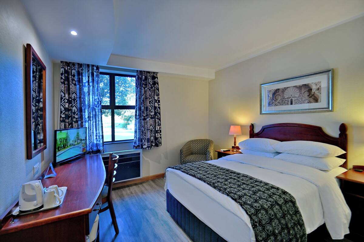 Une chambre d’hôtel avec un lit, un bureau équipé d’un ordinateur, une chaise, une fenêtre avec des rideaux et un tableau encadré au mur.