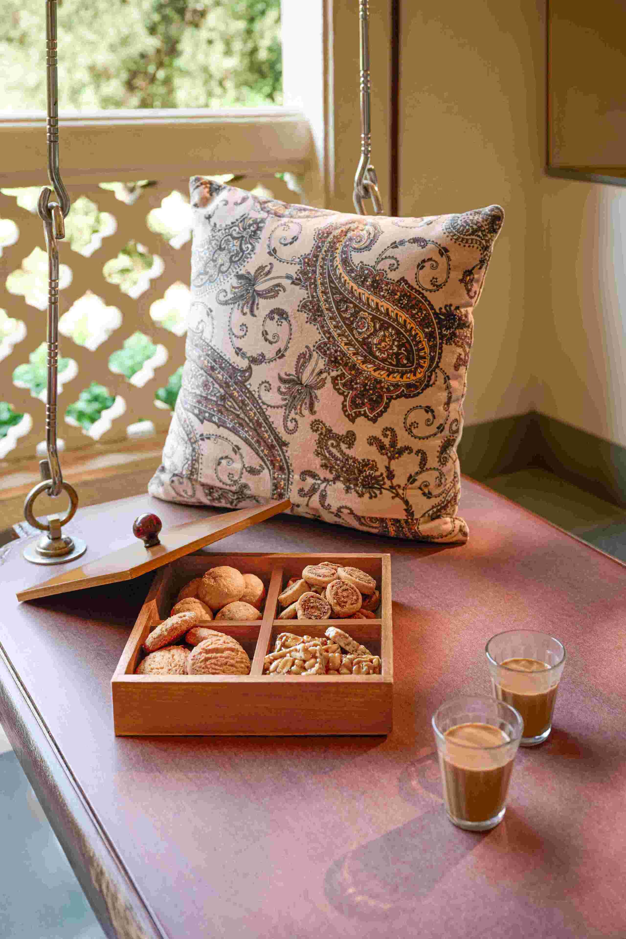 A wooden bench with a cushion, a tray of pastries, and two glasses of a brown beverage.
