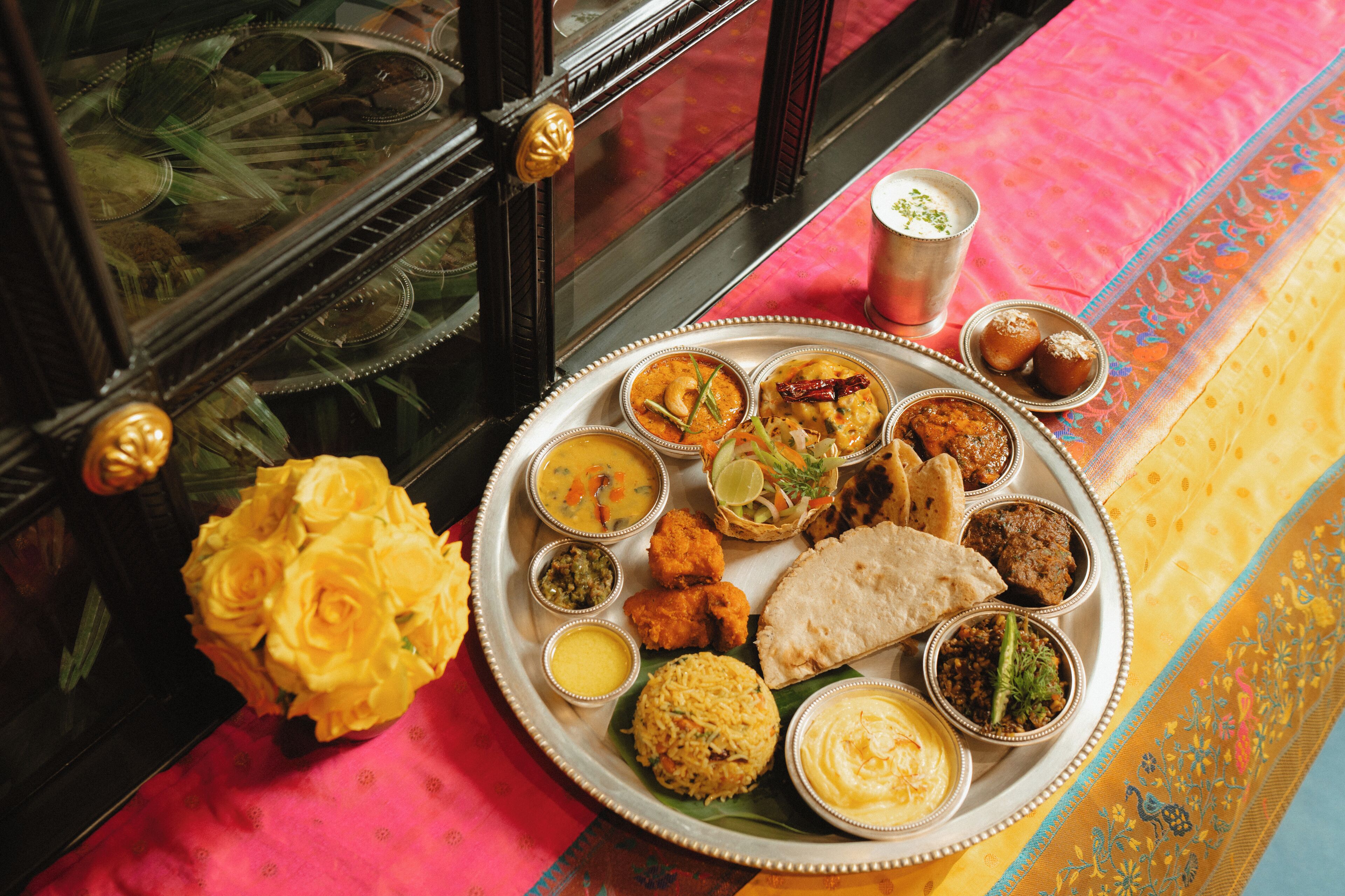 Una comida tradicional india con diversos platos sobre un platón de plata, incluyendo arroz, curries y pan, sobre un mantel colorido.
