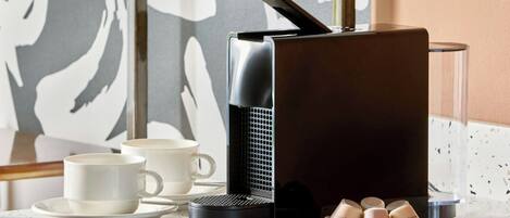 Una cafetera, una taza y un tazón pequeño con cápsulas de café sobre una encimera.