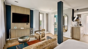 A modern hotel room with a large bed, a seating area with a sofa and armchair, a small table, a television mounted on the wall, a mirror, and a kitchenette. - The Deutz, a Tribute Portfolio Hotel by Marriott (Cologne)