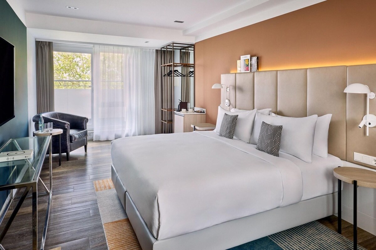 A modern hotel room with a large bed, a desk, a chair, a TV, and a wardrobe.
