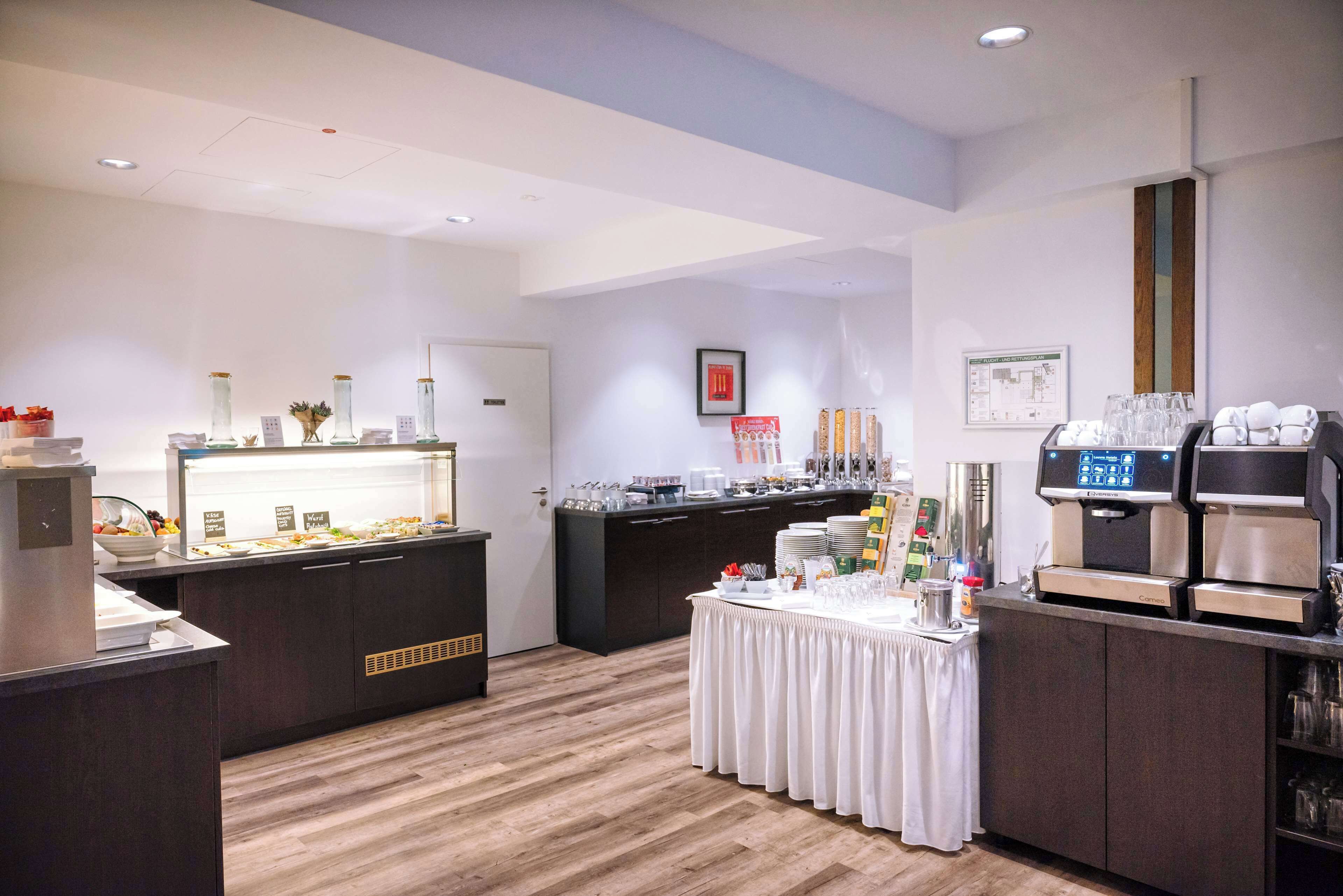 A modern hotel room with a wooden floor, a buffet area with various food items, a coffee station, and a small table with a white tablecloth.