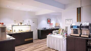 A modern hotel room with a wooden floor, a buffet area with various food items, a coffee station, and a small table with a white tablecloth.