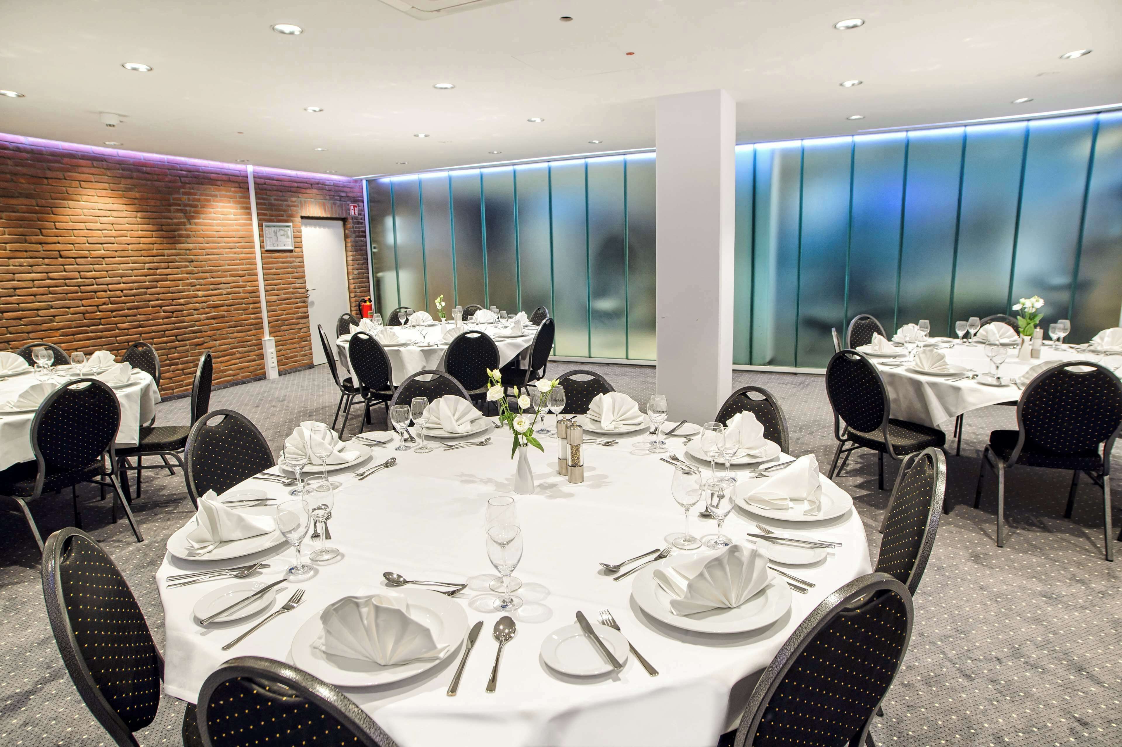 Un salón de conferencias con mesas redondas dispuestas para comer, equipado con manteles blancos, platos, cubiertos y copas.