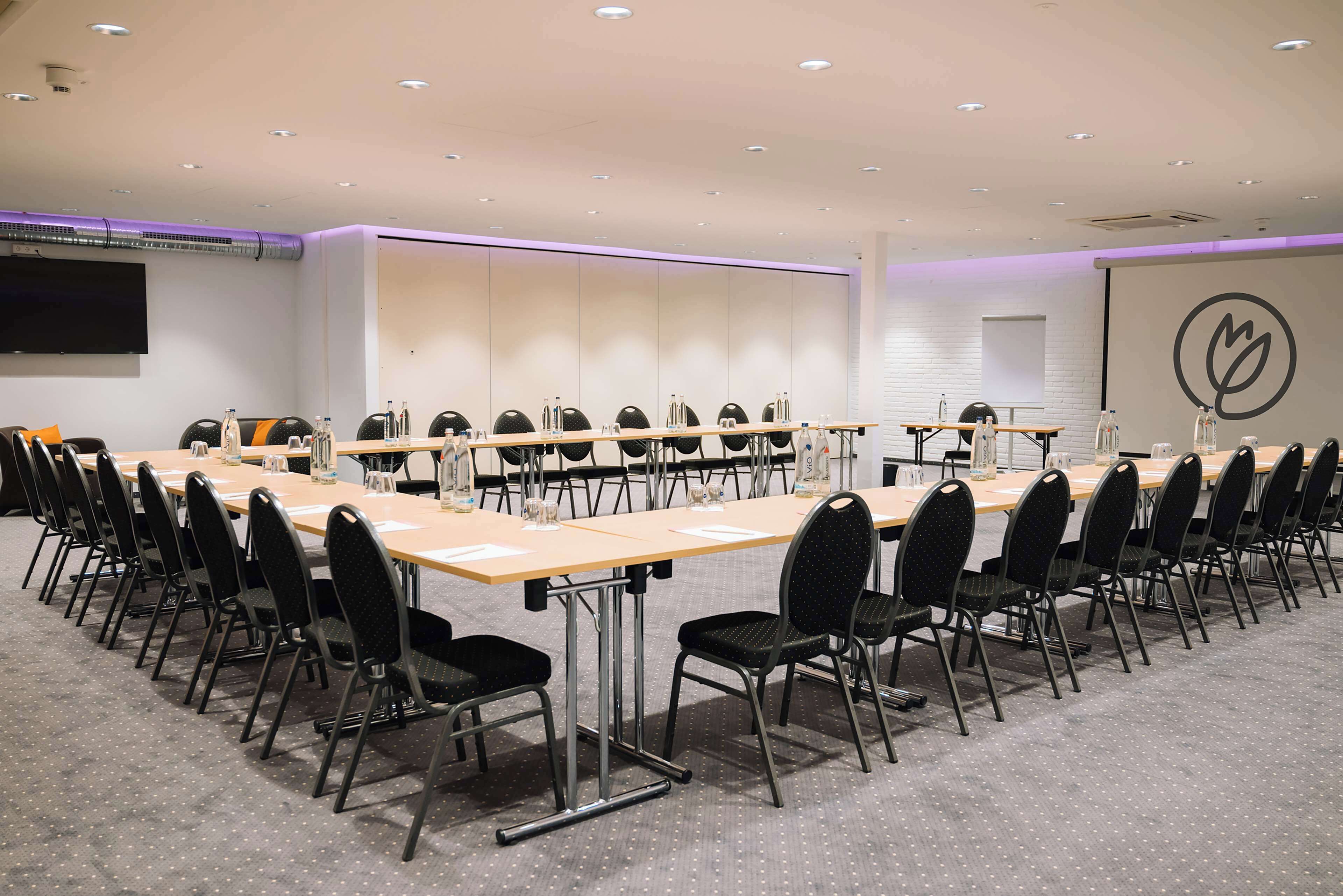 A conference room with a long table, black chairs, and a large screen on the wall.