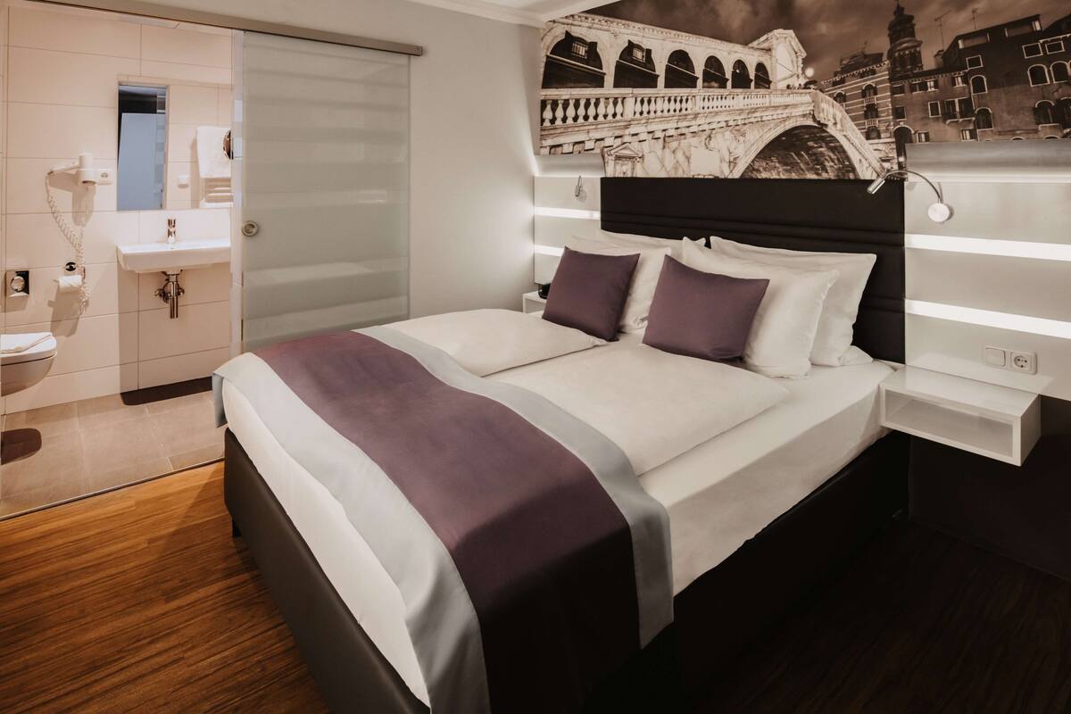 A modern hotel room with a large bed, a nightstand, a wall-mounted lamp, and a bathroom with a mirror, sink, and towel rack.