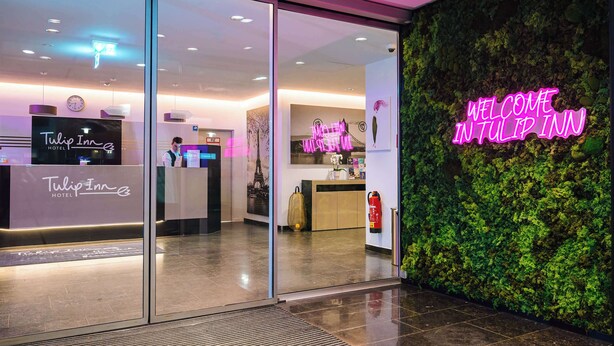 The entrance of Tulip Inn Hotel with a reception desk, a neon welcome sign, and a green wall with foliage.