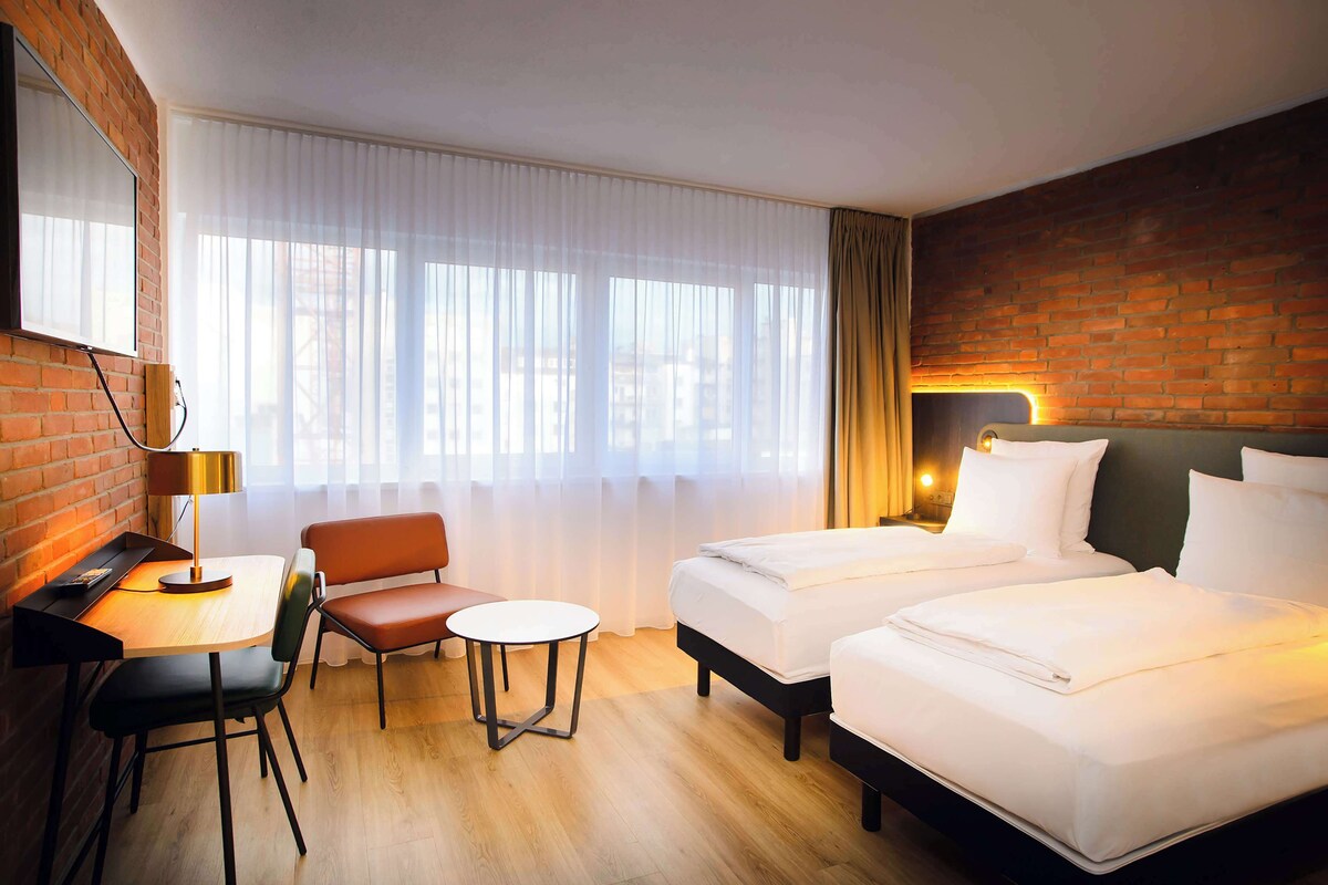 Une chambre d’hôtel avec deux lits, un bureau avec une chaise, une petite table et une vue sur la ville grâce à de grandes fenêtres.