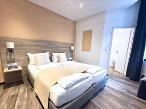 A modern hotel room with a large bed, bedside tables, a nightstand, a wall-mounted lamp, and a door leading to another room. - Hotel Hohenstaufen (Koblenz)