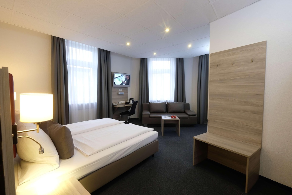 A hotel room with a bed, a desk, a sofa, a TV, and a wooden panel on the wall.