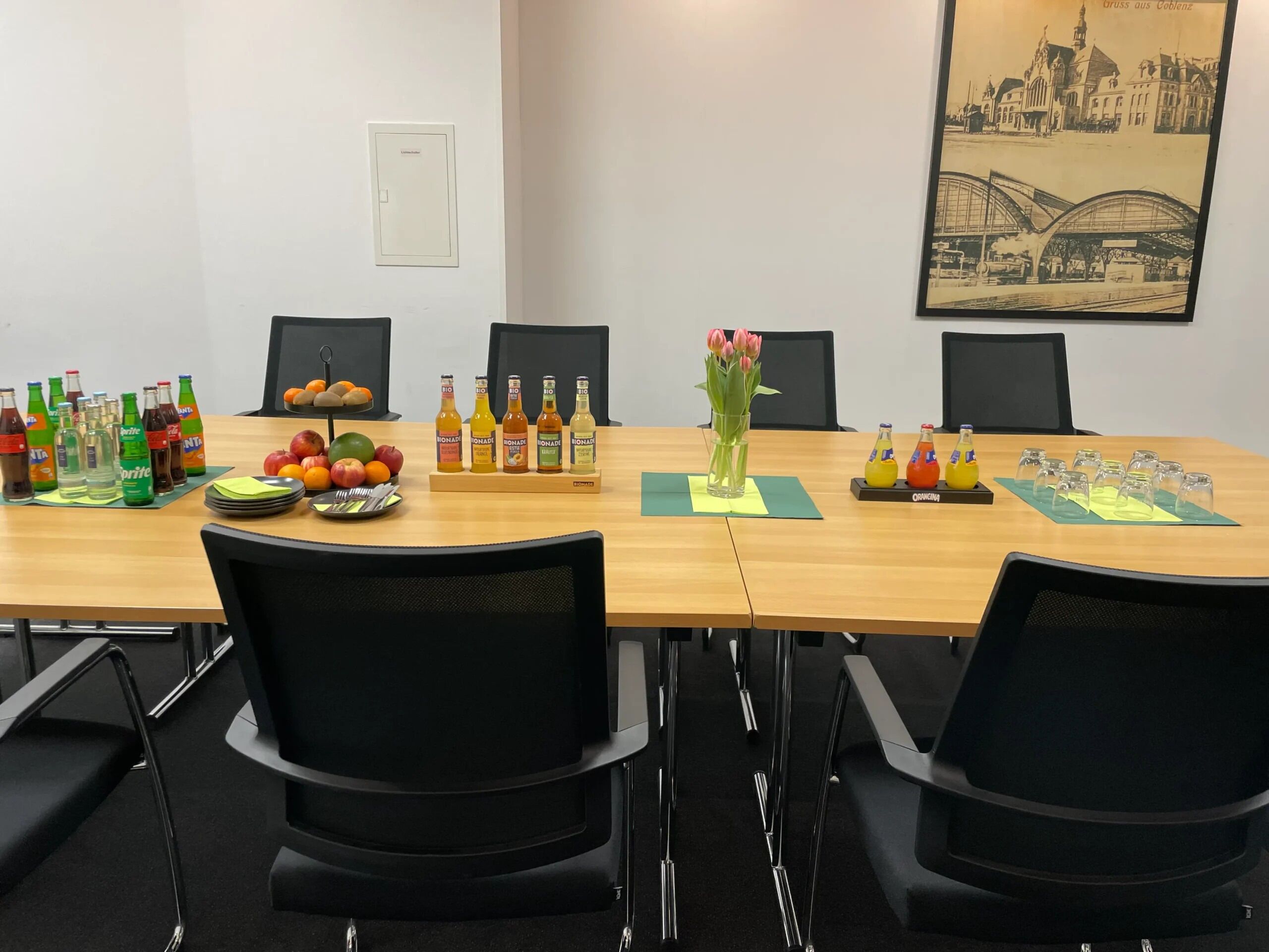 A conference room with a long table, chairs, a framed picture on the wall, and various items on the table including bottles and fruits.