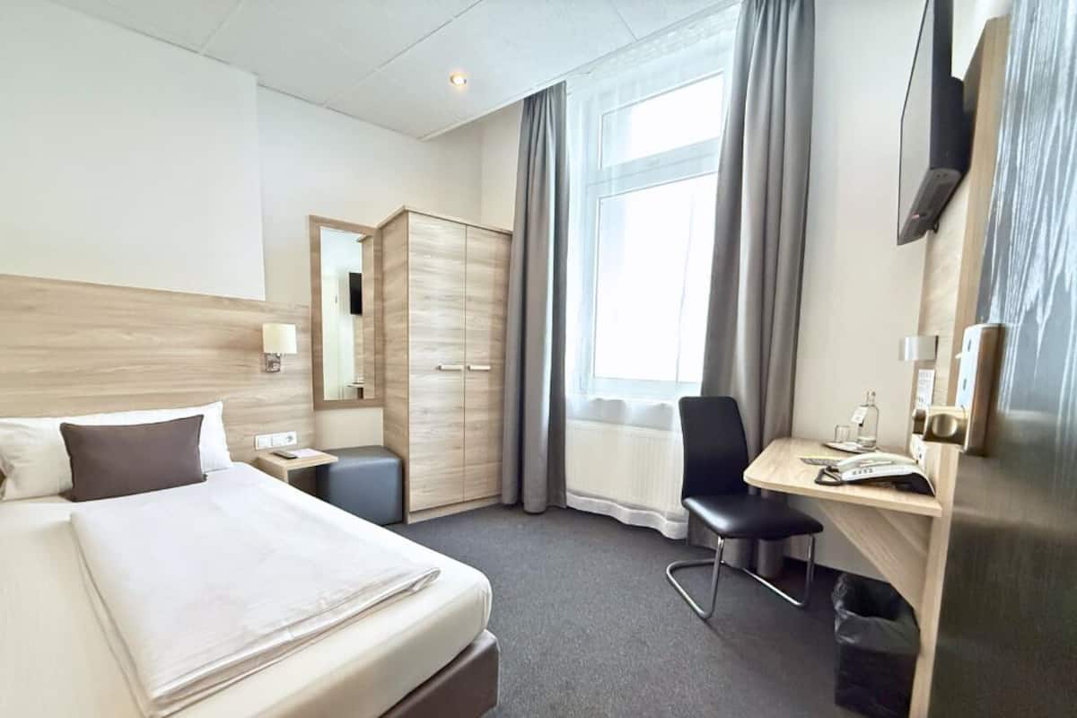 A hotel room with a bed, a desk, a chair, a wardrobe, a mirror, and a window with curtains.