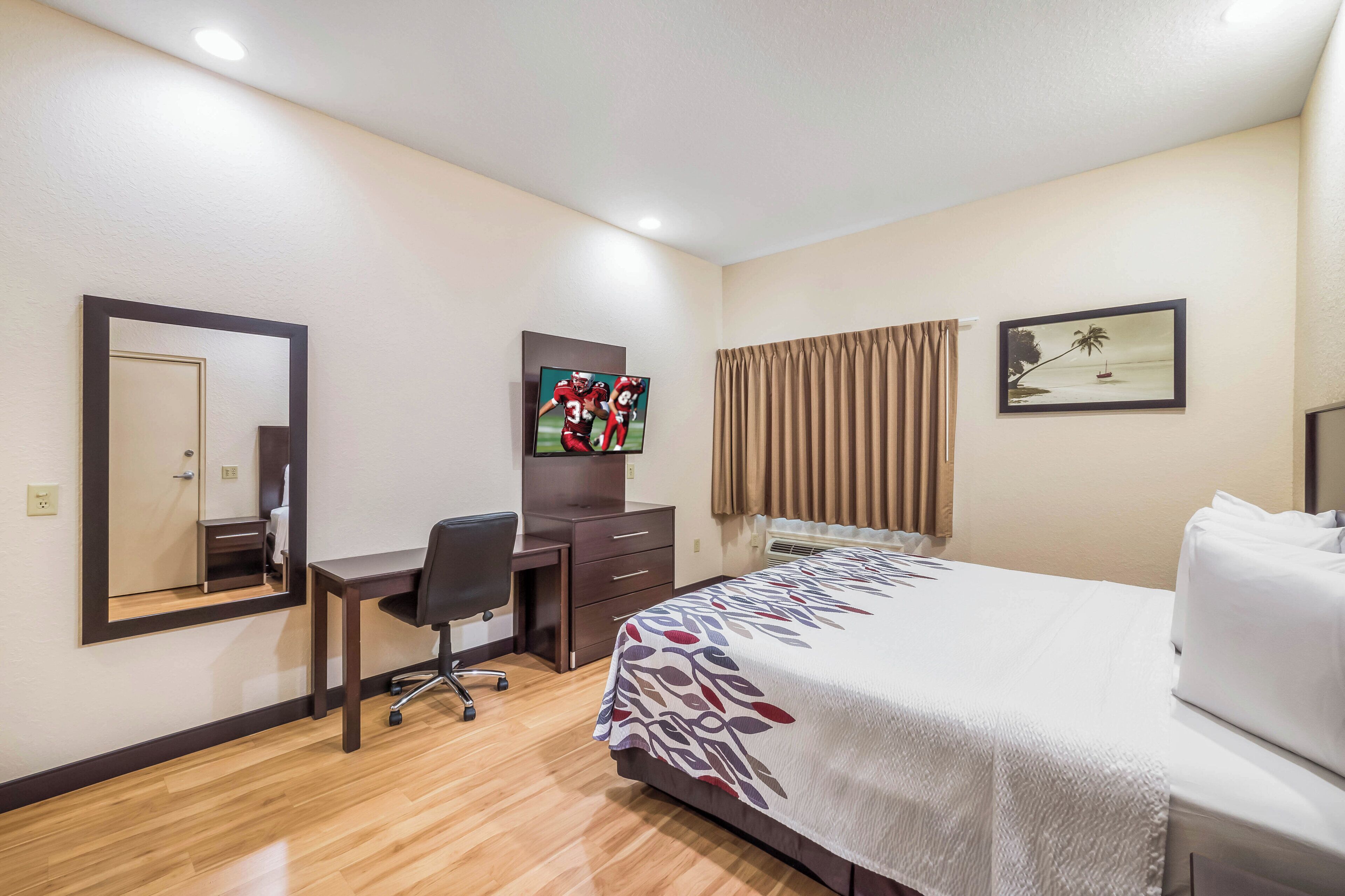 A hotel room with a bed, a desk, a chair, a television, a mirror, and a window with curtains.
