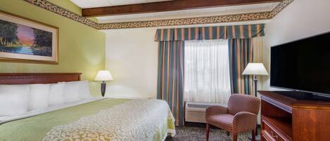 A hotel room with a large bed, two chairs, a TV, and a window with curtains.
