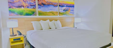 A neatly made bed with white bedding, a wooden headboard, and two bedside lamps. There are two paintings above the bed depicting coastal scenes.