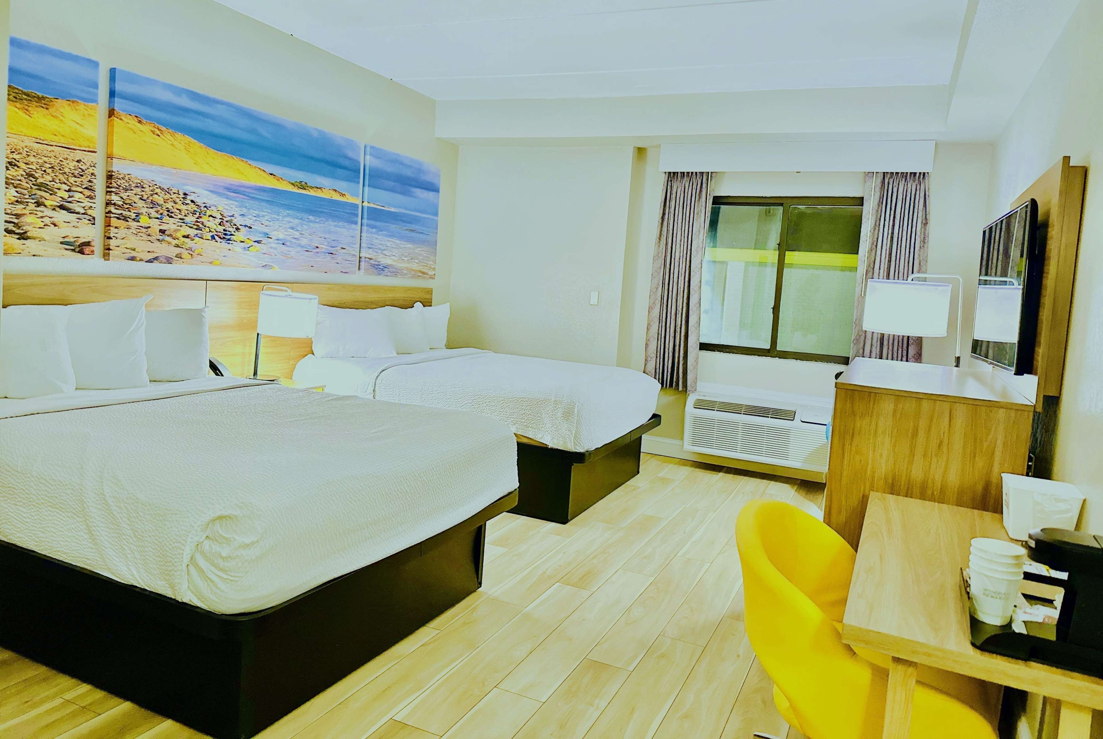 A hotel room with two beds, a desk with a chair, a large window with curtains, and a painting of a beach scene.