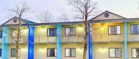 A multi-story motel with a yellow facade and blue accents, featuring balconies and a parking lot in front.