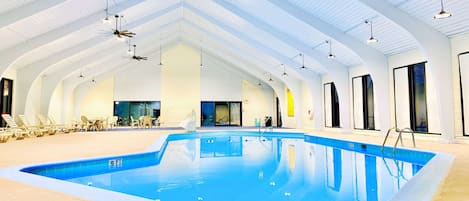 An indoor swimming pool with a clear blue water, surrounded by lounge chairs and a ceiling with white beams and hanging lights.