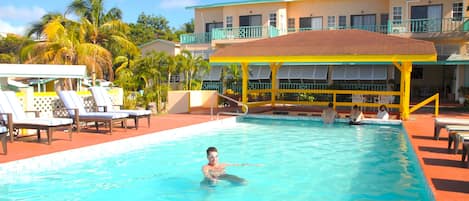 A swimming pool with lounge chairs and a covered area, surrounded by a building with balconies.