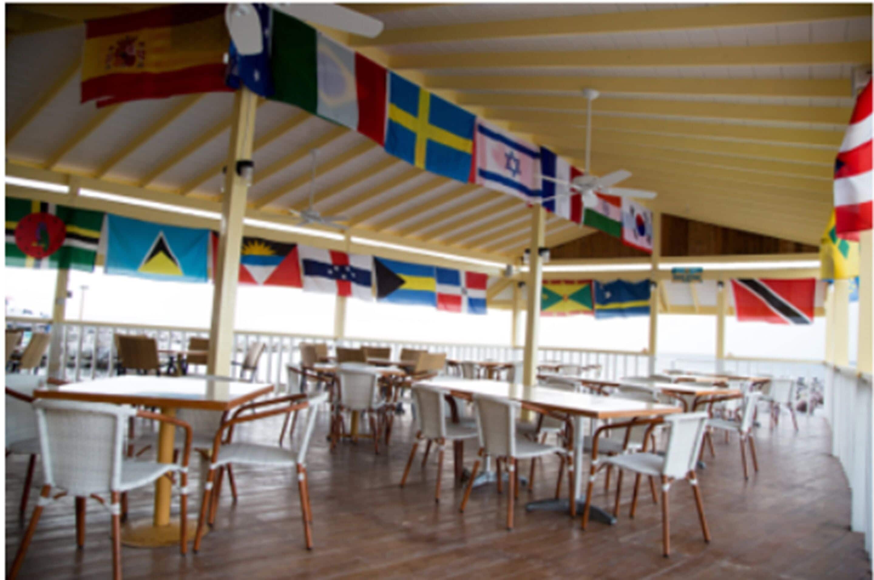 A covered outdoor dining area with tables and chairs, flags hanging from the ceiling, and a view of the sea.