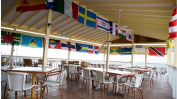 A covered outdoor dining area with tables and chairs, flags hanging from the ceiling, and a view of the sea.