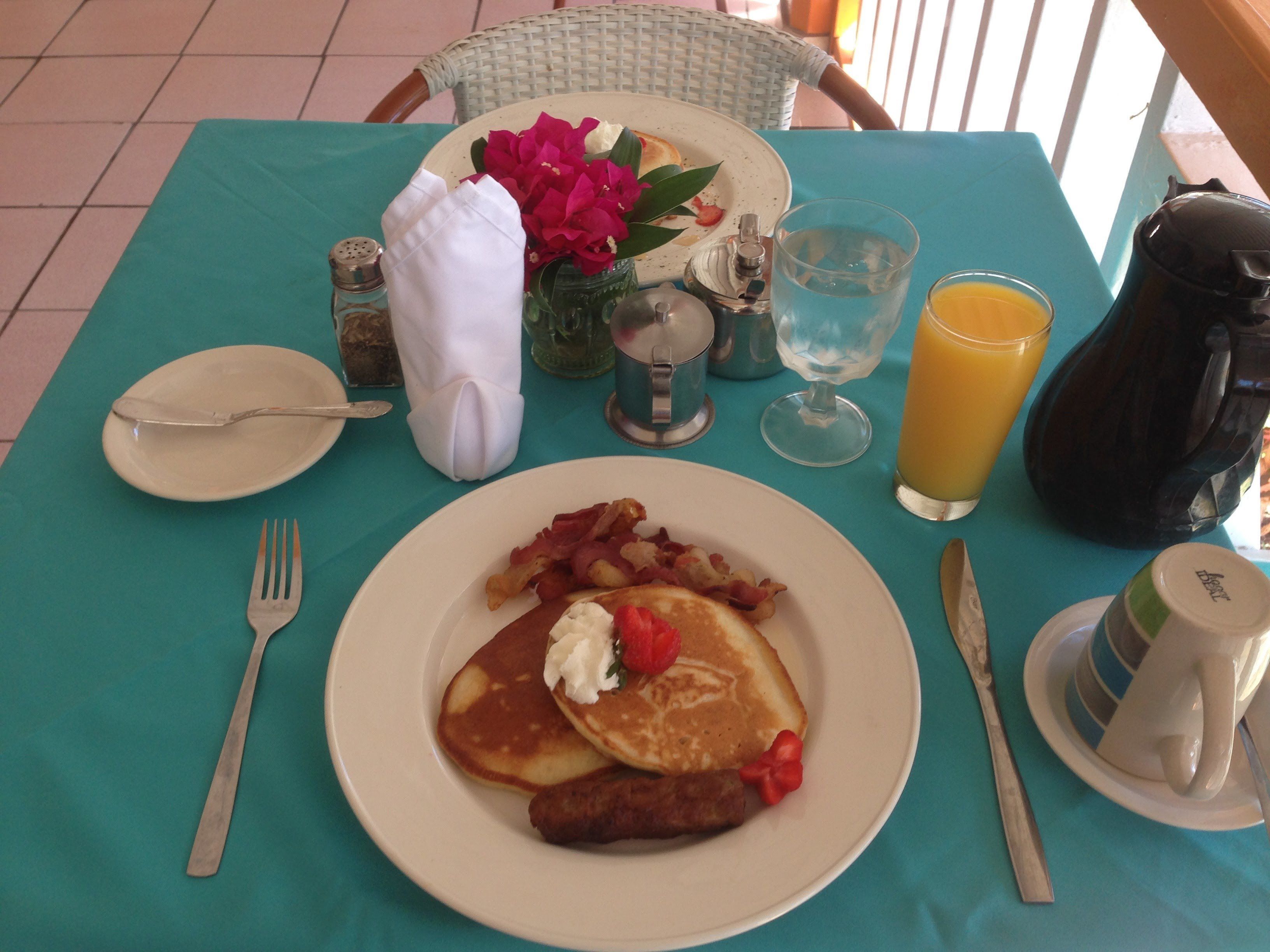 Uma mesa de café da manhã com panquecas, bacon e salsicha, um copo de suco de laranja e uma jarra de água.