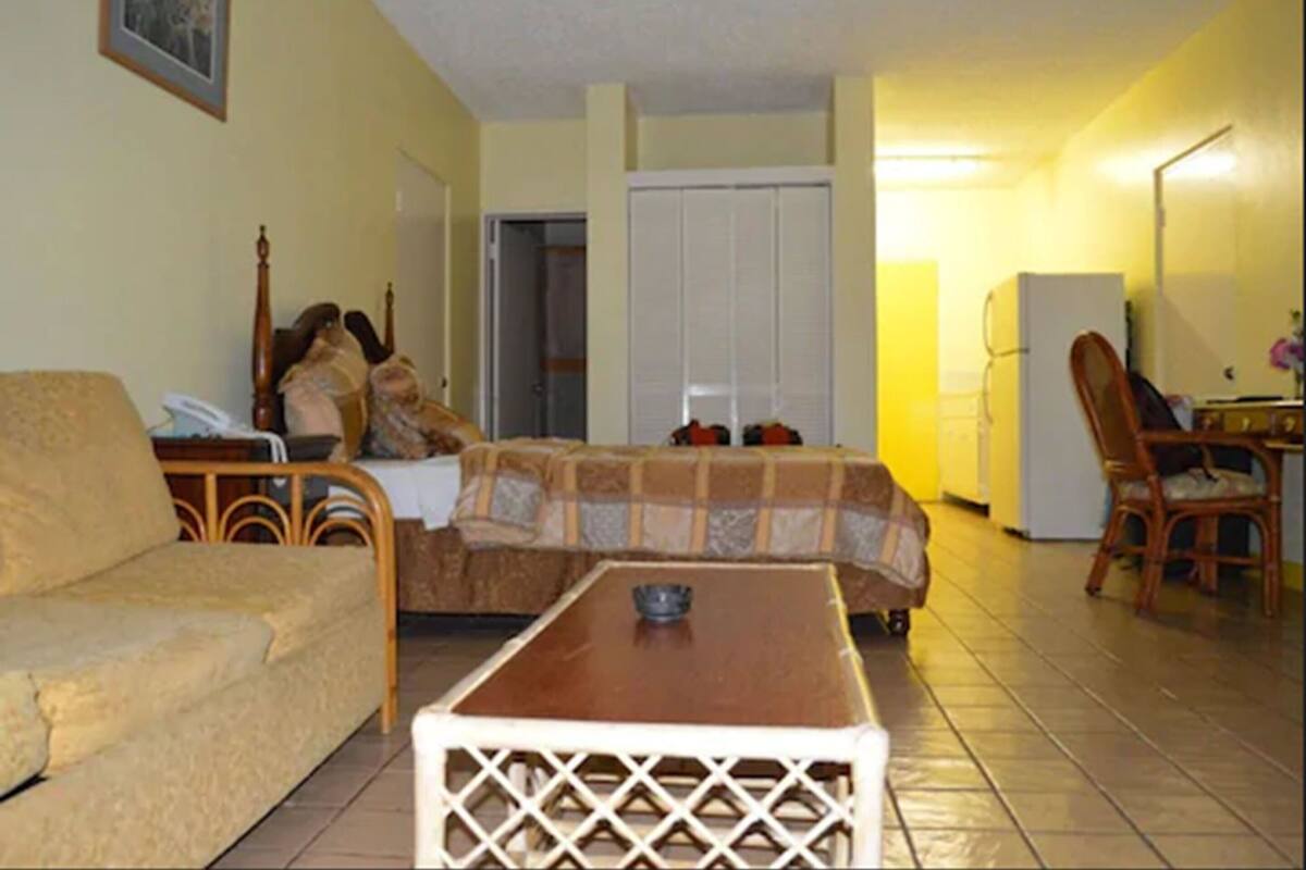 A living room with a sofa, a coffee table, a bed with a blanket, a wardrobe, and a refrigerator.