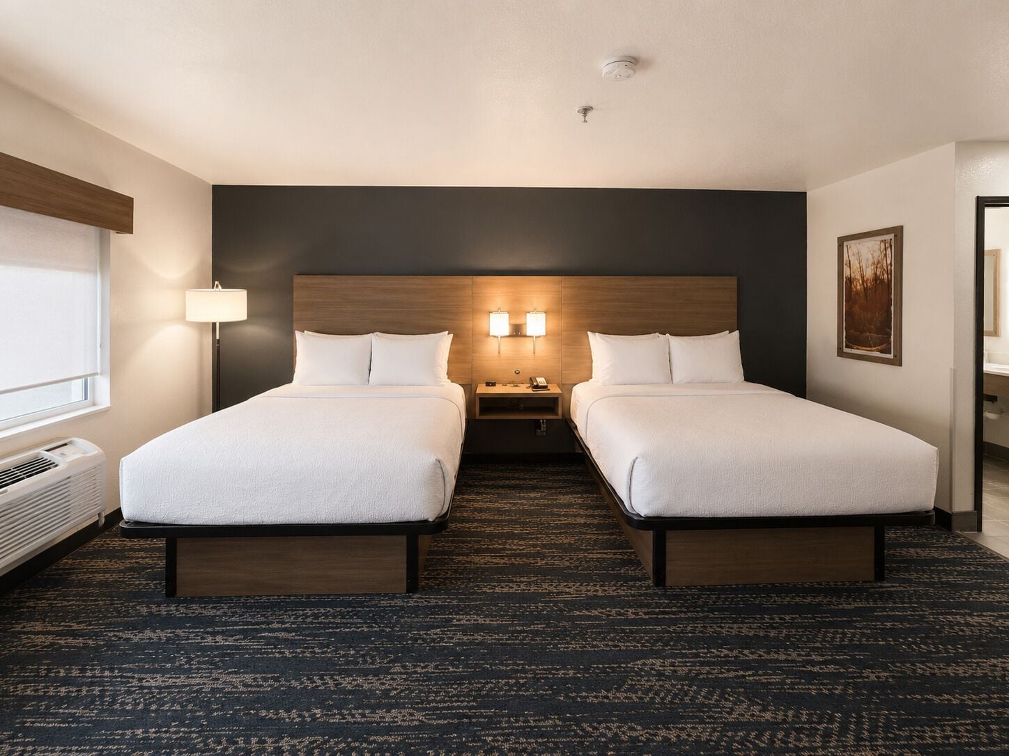 Una habitación de hotel con dos camas, una mesita de noche, una lámpara, un cuadro y una ventana.