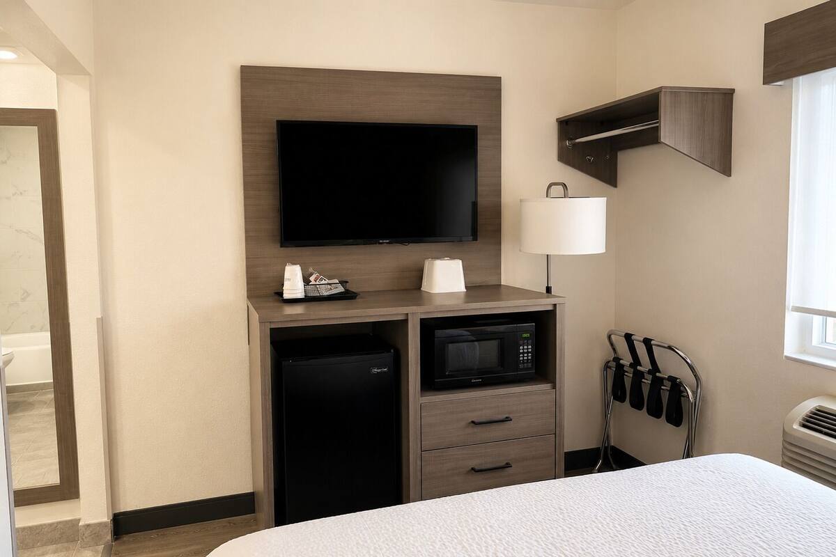 A hotel room with a bed, a flat-screen TV, a microwave, a mini-fridge, a coffee maker, a lamp, and a towel rack.