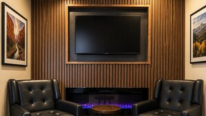 Una sala moderna con un televisor de pantalla plana montado en una pared de madera, dos sillones de cuero, una pequeña mesa redonda y una chimenea con iluminación azul.