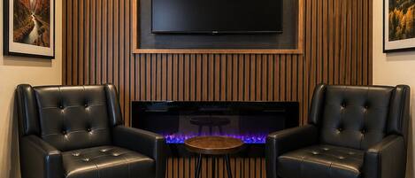 Una sala moderna con un televisor de pantalla plana montado en una pared de madera, dos sillones de cuero, una pequeña mesa redonda y una chimenea con iluminación azul.
